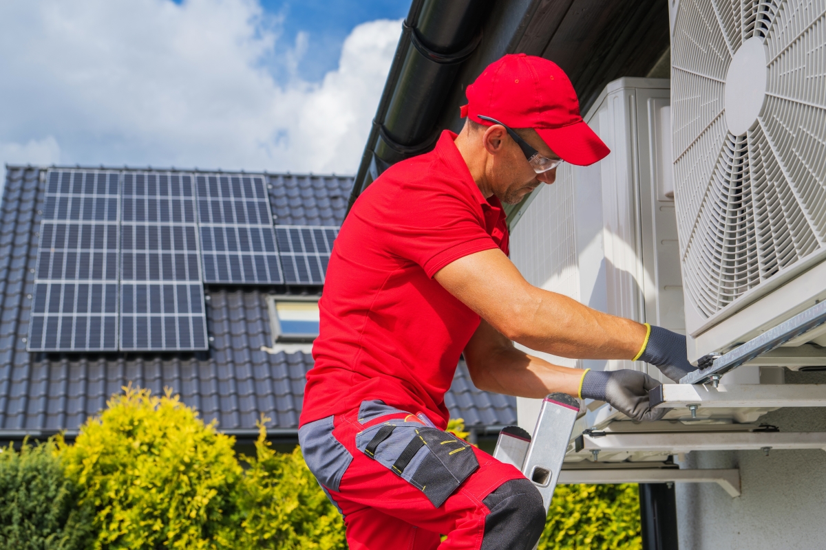 un artisan posant une pompe à chaleur sur un logement, panneaux solaires sur un toit en arrière plan