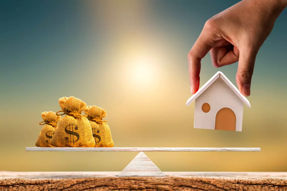 une balance en bois avec des sacs de dollars à gauche et une main qui s’apprête à poser une maison en bois à droite