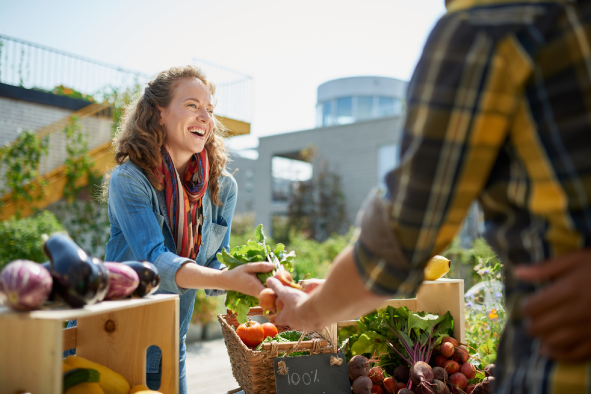  une femme maraicher tend des légumes à une main d’homme sur fond de jardin urbain