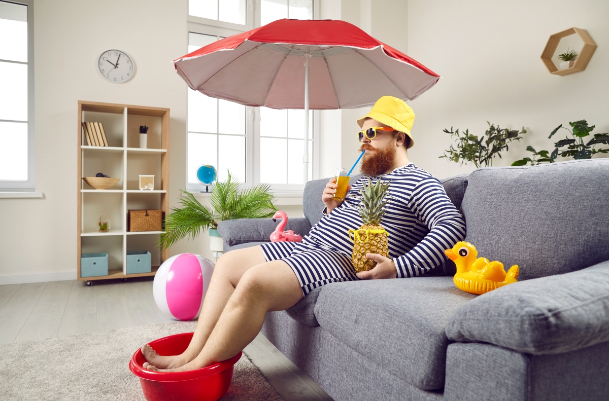 un homme vétu en maillot de bain couvrant à rayures sirote un verre avec une paille sous un bob et un parasol avec un ananas et un canard de bain, au milieu de son salon