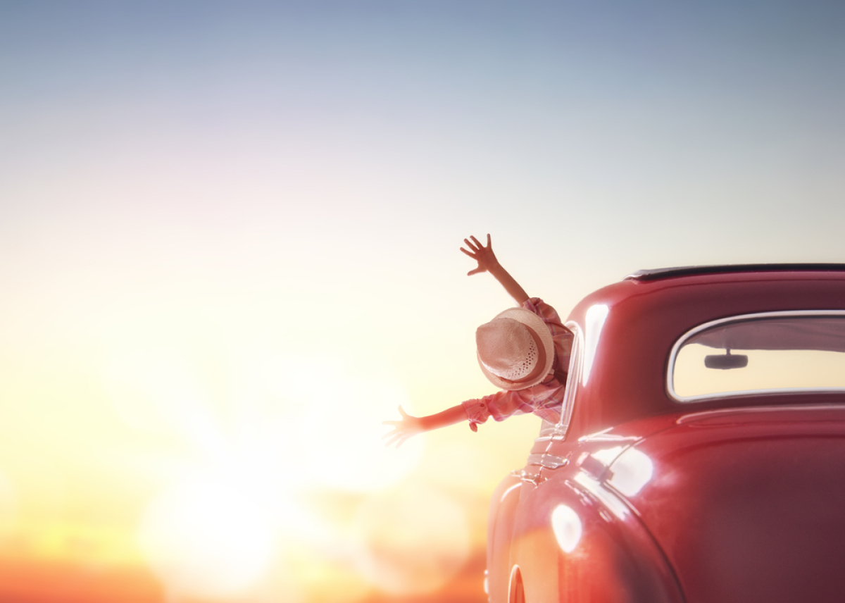 une femme avec un chapeau sort la tête et les deux ras par la fenêtre de sa voiture rouge au couchant