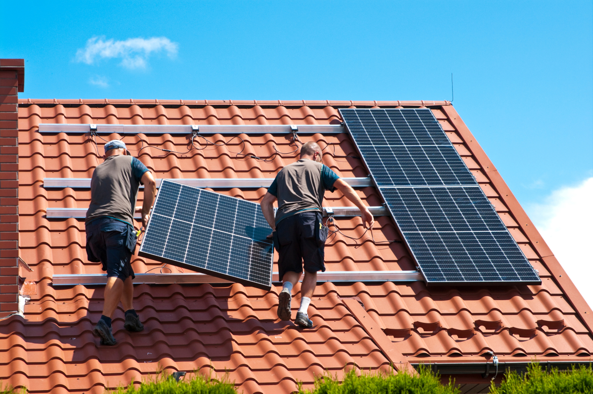 deux ouvriers installent des panneaux solaires sur un toit de tuiles rouges