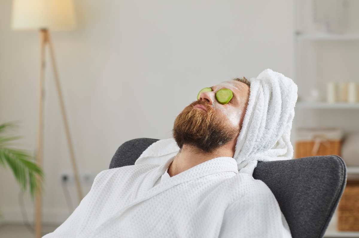 un homme barbu se détend sur un fauteuil avec une serviette en turban sur la tête et des rondelles de concombre sur les yeux